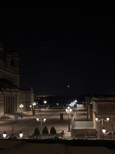 The image depicts a night scene of the Plaza de la Armería in front of the Royal Palace of Madrid. The plaza is illuminated by ornate streetlights. In the foreground, three conical-shaped trees are visible. To the left, the imposing facade of the Royal Palace is partially visible, characterized by its classical architecture with columns and windows. In the middle ground, the vast, paved plaza stretches out, with several streetlights casting pools of light. A few indistinct figures can be seen walking across the plaza. In the background, the cityscape of Madrid is visible, with a scattering of lights indicating urban development. The sky is dark, with a sliver of a crescent moon visible towards the upper right of the frame. The overall atmosphere is serene and nocturnal.