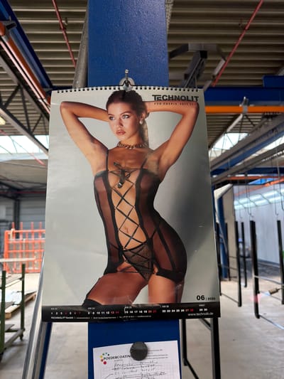 The image shows a calendar page from Technolit, featuring a woman in lingerie. The calendar is dated June 2026. The woman is leaning on her arm, looking towards the viewer. She is wearing a black, sheer lingerie set with criss-cross straps and a bow detail. The background of the calendar page is a light gray.
The calendar is attached to a spiral binding and is part of a larger print. There is visible text on the calendar page including the company name "TECHNOLIT", a tagline "FÜR PROFIS. SEIT 197", week numbers (KW 23-27), dates, and website address "www.technolit.de".
The calendar is placed in what appears to be an industrial setting. In the background, there are corrugated metal walls and metal structures, suggesting a factory or warehouse. There are also some red and orange pipes or conduits visible. A blue surface is in the foreground on the left, and part of another industrial structure is visible on the far right. The lighting suggests an indoor setting during the day. The overall impression is of a promotional calendar in a utilitarian environment.