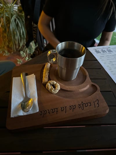A coffee cup is on a wooden tray with cookies and a napkin with a spoon. The tray is labeled "El café de la tarde". The scene is situated at El Oasis Valsequillo in Mexico. A person's arm, shoulder, and bag are visible, indicating someone is present at the table. There is a menu on the table next to the coffee cup and the tray. There is vegetation at the bottom of the image.