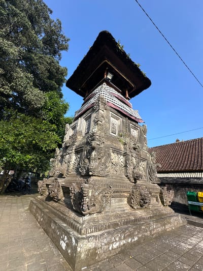 The image shows a detailed, multi-tiered stone structure with intricate carvings, topped with a thatched roof. The structure appears to be a temple or monument, possibly located in a tropical environment given the presence of lush green trees and a clear blue sky. The stone is weathered, showing patches of moss and lichen, indicating age and exposure to the elements. The lower section of the structure is a substantial stone base with decorative elements, possibly depicting mythological creatures. A black and white checked cloth is draped around the upper part of the stone section, just below the thatched roof. The roof itself is dark, possibly made of straw or reeds, and has some vegetation growing on it. In the background, there is a tiled roof of a building, suggesting an inhabited area. To the left of the structure, there are motorcycles parked, and to the right, two recycling bins, one green and one yellow, are visible. The ground is paved with stone tiles. The scene is well-lit by natural sunlight, casting shadows on the structure and pavement. The location is identified as Bangli, Indonesia.