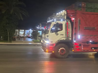 A white truck with a red cargo bed is driving on a wet road at night. The truck has many lights, including headlights, side lights, and roof lights. The cargo bed is loaded with stacked items, some of which are green and blue. In the background, there are palm trees and a building with lights on. The scene appears to be in Berrechid, Morocco, based on the context provided, likely on a main road with a nearby establishment. The road surface shows reflections from the truck's lights, indicating it might have rained recently.