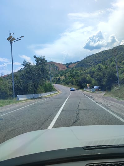 The image shows a view from inside a vehicle looking out at a winding road in a mountainous, rural area. A white car is driving on the road ahead, and another white car is parked on the right side of the road, near a small yellow structure. The landscape is covered in green trees and vegetation, with some barren, reddish-brown hills in the background. The sky is blue with scattered white clouds. There are power lines and streetlights visible along the road. The vehicle's hood is visible in the foreground of the image. The scene appears to be a sunny day. The location of Yusuphana, Uzbekistan is not depicted or indicated in the image.