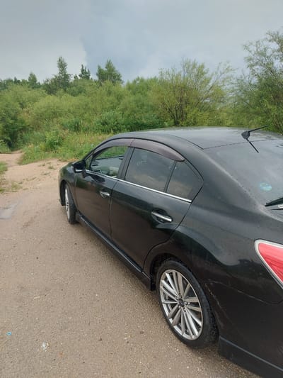 A black Subaru sedan is parked on a dirt road in Parunovka, Russia, surrounded by lush green foliage. The car is viewed from the rear passenger side, and its glossy black paint is speckled with raindrops. The road appears to be a path leading through a wooded area, with a small puddle of water visible on the left side of the frame. The sky is overcast, suggesting a cloudy or rainy day. The environment is natural and rural, with no visible buildings or other man-made structures in the immediate vicinity.