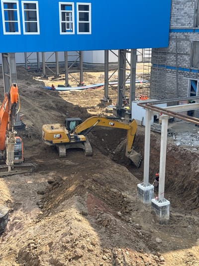 The image depicts an active construction site in Chulman, Russia, featuring heavy machinery and ongoing work. A large blue building with white-framed windows is visible in the upper portion of the frame. In the foreground, an excavator with the "CAT" logo is digging a trench. Another orange excavator, with a jackhammer attachment, is positioned to the left. Several workers in orange vests and hard hats are present on the site, actively engaged in construction activities. The ground is uneven and consists of dirt and rocks, indicating excavation and earthmoving. Concrete pillars supporting an overhead structure are also prominent in the scene, with ongoing work around their bases. The overall environment suggests a large-scale industrial or commercial construction project.