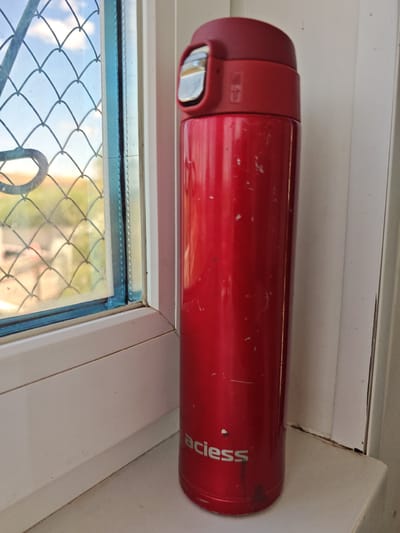 A red stainless steel thermos is standing on a white windowsill. The thermos has a silver button on the lid. To the left of the thermos is a window with a diamond-shaped grate. Outside the window, it appears to be daytime, with a blue sky and some greenery visible. The scene is located in the city of Prirechnoye, Kazakhstan.