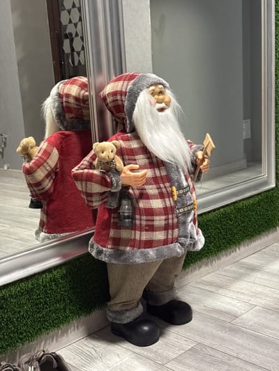 The image shows a Santa Claus doll leaning against a reflective surface. The doll is dressed in a red and beige plaid coat with grey faux fur trim. It has a long white beard and is holding a small teddy bear in one hand and what appears to be a wooden paddle in the other. It is wearing brown pants and black boots. The doll is positioned on a surface next to a strip of artificial grass. The background appears to be a reflective wall, possibly a display case or window. The location is not discernible from the image.