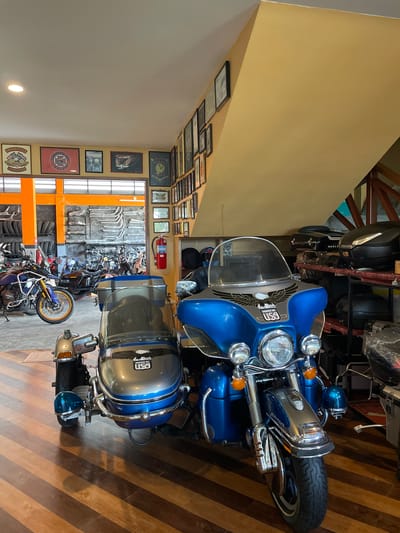This image depicts the interior of a motorcycle shop located in Ban Yid, Thailand. The foreground is dominated by a large, blue touring motorcycle equipped with a silver and blue sidecar, parked on a multi-toned wooden floor. The motorcycle features a clear windshield, chrome accents, and an eagle graphic with "MADE IN USA" text on its fairing; the sidecar also displays "MADE IN USA."

To the left, a yellow wall showcases multiple framed motorcycle club patches and emblems, including "IMMORTALS THAILAND" and "OLDTIMER MC THAILAND." The wall on the right, also yellow, is densely covered with numerous smaller framed pictures, extending up a sloped ceiling section. Below these frames, a shelving unit stores black motorcycle top cases and various accessories.

Through a wide, orange-framed opening in the midground, a workshop or storage area is visible. This back area features stacks of motorcycle tires on the left and a wall extensively covered with rows of chrome motorcycle exhaust pipes and other components. Several additional motorcycles are parked within this rear section, and a red fire extinguisher is mounted on the right wall. The ceiling is white with recessed lighting. No individuals are present in the visible scene.