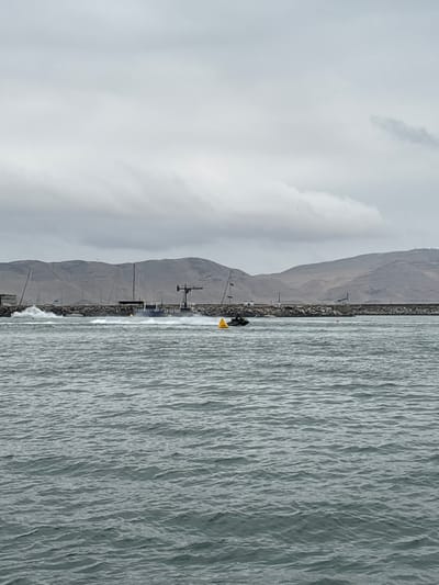 A jet ski is navigating a choppy body of water in front of a rock wall breakwater. The breakwater is lined with several sailboats with tall masts, and a large crane-like structure stands on the breakwater. Behind the breakwater, a range of barren, brown hills under a cloudy sky forms the background. The water is a deep gray-blue, with whitecaps indicating rough conditions. A yellow buoy is visible in the water to the right of the jet ski. The scene appears to be a harbor or marina area.
