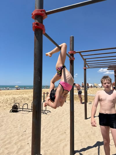 A young person is upside down on horizontal bars at a beach in Natukhayevskaya, Russia. The person is wearing a leopard print and pink bikini and a black cap. Another young person, a boy, stands to the right of the bars, facing the camera. In the background, the sea, beach, and other people can be seen. The weather is sunny with a clear blue sky.