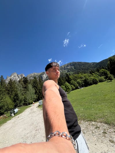 The image is a selfie taken outdoors in a mountainous, grassy environment under a clear blue sky with scattered clouds. In the foreground, a young man with short dark hair is captured from the chest up, wearing a black tank top and a silver chain bracelet on his left wrist. He is looking towards the right side of the frame. Behind him, a gravel path stretches into the distance. To the left of the path, large, rocky mountains are visible, interspersed with dense green pine trees. To the right of the path, a lush green meadow slopes upwards towards a forested mountainside. A person in red shorts is walking away from the camera on the path in the middle ground. Two large, cylindrical metal containers, possibly for waste disposal, are situated beside the path. The overall scene suggests a rural or natural setting, potentially near a hiking trail or a natural park. The location is identified as Govedartsi, Bulgaria.