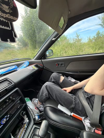 The image shows the interior of a car, looking towards the passenger side. A person is seated in the passenger seat, wearing ripped black jeans and grey sneakers. They have a seatbelt fastened. On the dashboard, two packages of Oreos are visible. A "Top Gear" air freshener hangs from the rearview mirror. The car appears to be driving through a rural or undeveloped area with trees and greenery visible through the windows. The sky is blue with some clouds. The location is identified as Kumachevo, Russia.