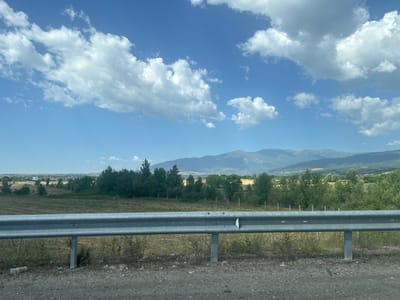 This is a landscape photograph taken from a roadside, likely from a vehicle, looking out over a rural area. The foreground features a grey metal guardrail, typical of a highway or main road, with a dirt and gravel shoulder. Beyond the guardrail, a dry, grassy field stretches out, with some sparse vegetation and evidence of agricultural activity. A line of trees forms a mid-ground boundary, and in the distance, rolling hills and mountains are visible under a bright, blue sky filled with scattered white clouds.
The scene suggests a sunny day, possibly late morning or early afternoon, given the lighting conditions and the clear sky. The location appears to be rural and possibly mountainous, consistent with the context of Barakovo, Bulgaria. There are no people visible in the image. There is some litter on the ground near the guardrail, including a plastic bottle. In the far distance, a small settlement or town can be seen nestled amongst the landscape. A billboard or large sign is visible on the left side of the horizon. The overall mood is peaceful and natural, characteristic of a scenic countryside view.