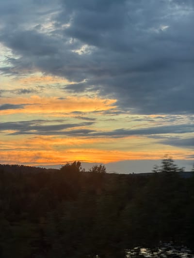 The image shows a landscape at sunset or sunrise, potentially located near Arsk, Russia. The sky dominates the scene, filled with elongated clouds that are illuminated by the light from the setting or rising sun. The color of the sky transitions from a deep grey with darker clouds to vibrant orange and yellow hues near the horizon. The horizon line is marked by a dark silhouette of trees and vegetation, suggesting a forested area or shoreline. The lower portion of the image appears to show water, possibly a lake or river, reflecting the colors of the sky. The overall impression is one of tranquility and natural beauty, capturing a fleeting moment of atmospheric light and shadow. The image is rotated 90 degrees counter-clockwise.