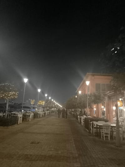 The image captures a vibrant nighttime scene of a pedestrian promenade in Palaio Faliro, a coastal suburb of Athens, Greece. A wide, paved walkway, appearing to be made of brick or stone pavers, stretches into the distance, illuminated by an array of lights.

On both sides of the promenade, numerous outdoor dining areas are set up with neatly arranged white tables and chairs, suggesting a lively row of restaurants, cafes, or bars. On the right, these establishments are housed within low-rise buildings characterized by a distinctive reddish-orange facade and large windows. Small, manicured trees line the walkway and stand in front of the buildings, many adorned with bright, decorative string lights that add a festive glow.

The entire area is well-lit by a succession of tall, modern streetlights emitting a clear white light, complemented by the softer, often yellow, glow from the decorative lights on the trees and buildings. In the middle distance, several indistinct figures can be seen walking casually along the promenade, indicating a moderate level of evening activity. The dark night sky looms above, providing a stark contrast to the brightly illuminated pathway below. The overall ambiance is inviting and relaxed, typical of an evening stroll or dining experience in a Mediterranean setting. No discernible text is visible in the image.