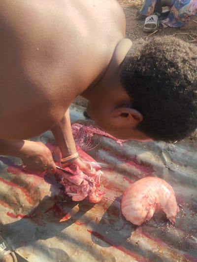 A man hunches over a blood-splattered surface, wielding a knife to cut raw meat. The scene suggests butchering or preparation of an animal. Various organs or meat parts are visible, including what appears to be a large stomach or organ sac. An Adidas sandal is visible in the upper right. The overall environment appears outdoors and the man is shirtless with dark skin.