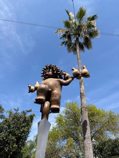 A statue of a boy with radiating hair holds chickens stacked on his head and arms. The sculpture sits atop a pole anchored by a vertical rectangular shape. A tall palm tree stands to the left of the statue with a clear blue sky visible behind. Green trees and foliage surround the base of the statue. The image is taken from a low angle, looking up at the statue and trees. Power lines run across the sky in the upper left corner.