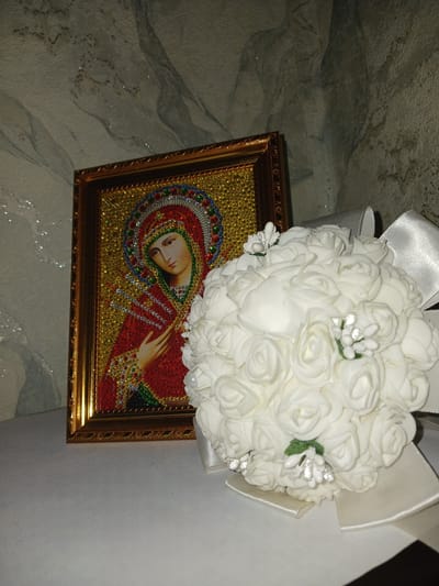 A framed religious icon depicting Mary is situated on a white surface next to a bouquet of white artificial roses. The icon is adorned with small, colorful gemstones and is framed in gold. The bouquet is large and round, with white ribbons tied around it. The background appears to be a textured wall. The scene suggests an indoor setting, possibly a home or a religious establishment. There are no people or animals visible in the image. The image is cropped from a wider scene, indicating that it is part of a larger collection of photographs. The location of Prokudskoye, Russia, is not discernible from the visual information provided in the image.