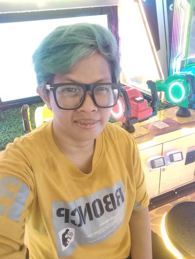 A middle-aged individual with blue-tinted hair and black-rimmed glasses is taking a selfie in an arcade. They are wearing a yellow t-shirt with blue graphic prints on the sleeves and front. Behind them are two arcade game machines with bright lights and colorful designs. The floor appears to be covered in artificial green turf. The overall environment suggests a recreational setting in Gunung Putri, Indonesia.