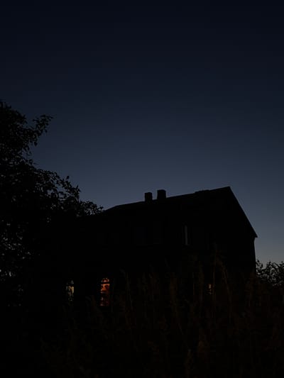 The image depicts a dark, silhouetted house with a peaked roof and two chimneys against a twilight sky. The sky is a deep, dark blue, transitioning to a lighter shade near the horizon. In the foreground, the dark shapes of trees and tall grasses obscure the lower portion of the house. Two windows on the side of the house are illuminated from within, casting a warm, orange glow. The overall atmosphere is somber and quiet, suggesting a time just after sunset. The location is identified as Leninsk-Kuznetskiy, Russia, but no specific landmarks or contextual details are visible in the image to confirm this.