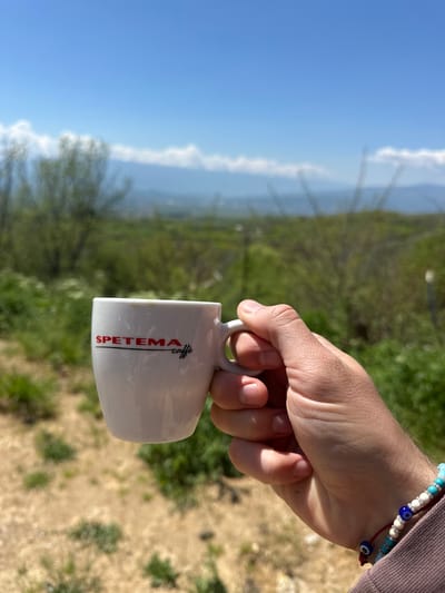 A person's hand holding a white coffee mug with "SPETEMA caffe" printed on it, with a landscape background. The foreground is dominated by the hand holding the mug. The wrist has two bracelets, one with turquoise, white, and blue beads, and another with a red cord and small blue beads.
The background appears to be a hilly landscape with vegetation, possibly trees and shrubs. In the distance, there's a hazy blue sky, suggesting a clear day. The angle of the photograph makes it difficult to determine the exact location. Based on the location you provided, the landscape aligns with the hilly terrain found in Bulgaria near Lyubovka. The image appears to have been taken outdoors in natural lighting.