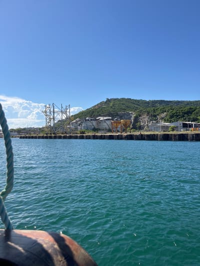 The image captures a vibrant, sunny coastal or lakeside scene, showcasing a blend of natural landscape and industrial structures. The photograph is taken from a low vantage point, likely from a boat or a dock.
In the immediate foreground, a sturdy, blue-green braided rope stretches horizontally across the bottom right of the frame. To its right, a dark, rounded object, possibly part of a boat's hull or a large wooden post, is partially visible.
The right side of the image is dominated by a wide expanse of water, which is a clear, deep blue-green color. The surface of the water is covered with numerous small ripples and waves, reflecting the bright sunlight and creating a shimmering effect.
On the left side of the image, a steep, rocky cliff face rises dramatically from the water's edge. The cliff is extensively covered with dense, green vegetation, primarily trees and shrubs, giving it a lush appearance. Along the base of this cliff, several man-made structures are evident, suggesting industrial or construction activity. These include what appear to be concrete structures, possibly buildings or foundations, and distinct yellow or orange crane-like frameworks. Further to the left along the shoreline, a large metal lattice structure, resembling a power transmission tower or a heavy industrial frame, stands tall against the backdrop of the cliff. The edge where the land meets the water appears to be a constructed pier or a fortified embankment.
The sky, occupying the upper portion of the image, is a brilliant, clear blue, indicating excellent weather conditions. A few puffy white cumulus clouds are visible on the horizon, above the industrial structures and the water, adding a touch of softness to the otherwise stark blue sky.
The overall impression is that of a bright, clear day, with strong sunlight illuminating the scene. The image presents a striking contrast between the verdant natural cliff and the utilitarian, man-made structures along its base, set against the calm, shimmering water. No visible text is present in the image.
