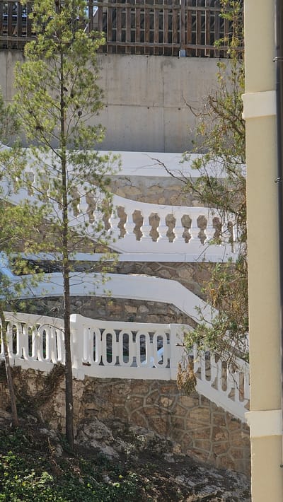This is a medium-shot, eye-level, outdoor photograph of a terraced staircase with white balustrades and stone retaining walls. The staircase is situated on a hillside with sparse vegetation, including pine trees and ivy. In the background, a concrete wall supports a wooden fence. The overall scene appears to be in a natural or landscaped setting within the city of Alhama De Aragón, Spain. There are no people or animals visible in the frame.