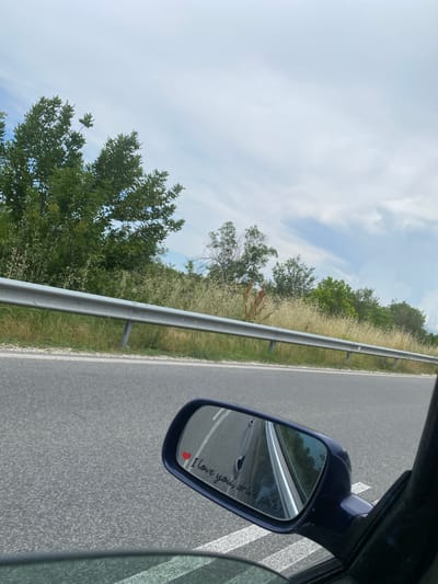 The image shows a view from a car window, looking out at a highway with a guardrail and vegetation along the side. In the foreground, a side-view mirror is visible, reflecting the road and the car's interior. On the mirror, a small red heart sticker is placed next to the handwritten text "I love you, drive safe". The sky above is partly cloudy. The scene does not depict any elements that would allow for identification of the specific location within the city of Aleko Konstantinovo, Bulgaria.