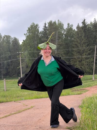 A woman stands on a dirt path under a rainy sky in what appears to be a rural area of Nizhnii Vishur, Russia. She is wearing a green shirt, black pants, and a black jacket. A large green leaf rests on her head. She is smiling. Trees and power lines are visible in the background. The ground is damp, and rain streaks are visible throughout the image.