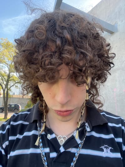 A medium-range, tilted, eye-level shot depicts a person with curly brown hair. Two braids are visible near the face, one on each side. The person's lips, nose, and part of the face are visible. The person is wearing a striped black and light blue polo shirt. Several necklaces are visible. The background includes a concrete structure and a tree. A car is visible in the distance. This image was taken in Bahía Blanca, Argentina.
