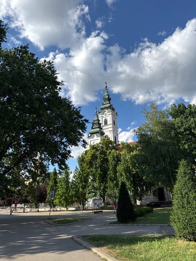 This image depicts the Serbian Orthodox Church of Saint George (Crkva Svetog Đorđa) situated in Becej, Becej Municipality, Serbia. The scene is captured on a bright day under a vivid blue sky interspersed with substantial white cumulus clouds.

The central subject is the white-painted church, characterized by two prominent bell towers featuring green, onion-domed spires, each surmounted by a cross. The tower on the right side of the image displays a visible clock face. The church's architecture appears to be in a Baroque style.

The structure is framed and partially obscured by a variety of mature green trees. A large, dense-foliaged deciduous tree occupies the left foreground and mid-ground, while a mix of deciduous and coniferous trees with lighter green foliage lines the right side and extends into the background, surrounding the church.

In the foreground and mid-ground, a paved pathway or road segment is visible, with distinct tree shadows cast upon its surface. Patches of green grass and low shrubs define the edges of the paved areas. To the left and in the background, a public area with benches is discernible, and faint human figures can be seen walking or seated. A white circular street sign marked "ЗОНА" (ZONE) featuring a pedestrian and bicycle pictogram is positioned on the left side of the pathway. Faint structures and fences are visible in the background through the trees.