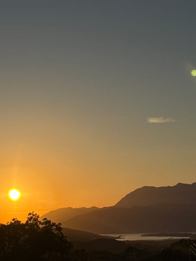 The image depicts a scenic sunset over a mountainous landscape in Prijeradi, Montenegro. The sun, a bright orb of yellow, is low on the horizon, casting a warm orange glow across the sky and illuminating the tops of distant mountains. Silhouetted trees in the foreground add depth to the scene. In the distance, a body of water, possibly a bay or a lake, is visible between the mountain ranges, with a few faint lights suggesting human habitation. The sky transitions from a pale orange near the horizon to a muted blue overhead. A faint, wispy cloud is present in the upper right quadrant of the sky, and a circular, greenish light source, likely a lens flare or reflection, is also visible in the upper right portion of the image. The overall impression is one of tranquility and natural beauty.