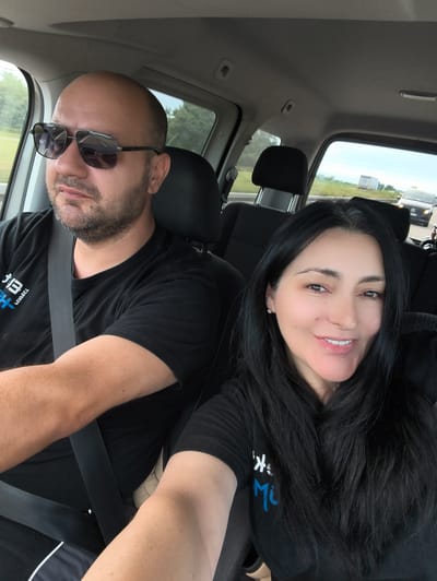 The image shows two people, a man and a woman, inside a vehicle. The man, on the left, is driving and wearing sunglasses and a black t-shirt. He has a beard and is looking to his left. The woman is in the passenger seat and is looking at the camera, smiling. She has long, dark hair and is wearing a black t-shirt. The interior of the vehicle is visible, including the dashboard, seats, and seatbelts. Through the windows, a road and trees can be seen, suggesting the vehicle is in motion. The sky appears overcast. The scene suggests a casual road trip. The question about the location in Hartheim am Rhein, Germany, cannot be answered from the visual information provided in the image.