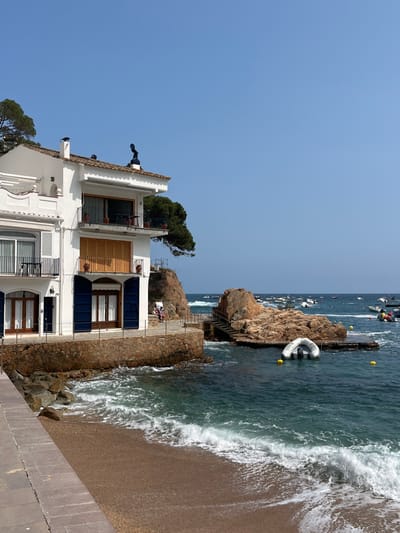 A white two-story building with a balcony and blue shutters sits on a rocky shore in Tamariu, Spain (latitude: 41.918, longitude: 3.208). Two people are sitting on a ledge in front of the building. A small white dinghy is moored in the water in front of the building. The water is a bright blue and the sky is clear. The beach is sandy and there are rocks in the foreground. The shore is lined with a low wall and there is a narrow paved walkway leading to the water. There are several small boats in the distance. The scene is peaceful and serene.