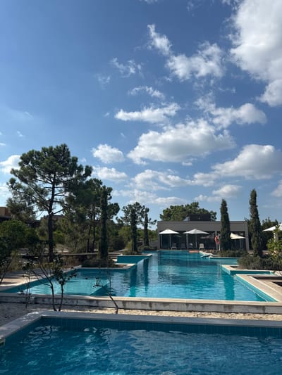 This image presents an outdoor swimming pool complex in Grândola, Portugal, captured under a bright blue sky with scattered white cumulus clouds. The foreground and midground are dominated by several interconnected swimming pools, distinguished by light blue tiling and clear, still water that reflects the sky. The pools feature light-colored coping and are separated by narrow sections of light-toned sand or gravel, interspersed with small, sparse vegetation. A red lifebuoy hangs on a pole near one of the larger pool sections, which also contains a light-colored inflatable float. In the background, a contemporary, flat-roofed building with large glass panels and a grey facade is visible, flanked by two white patio umbrellas. The entire complex is surrounded by mature trees, primarily tall pine trees and slender cypress-like trees, suggesting a natural or resort-like setting. No individuals are present in the scene.