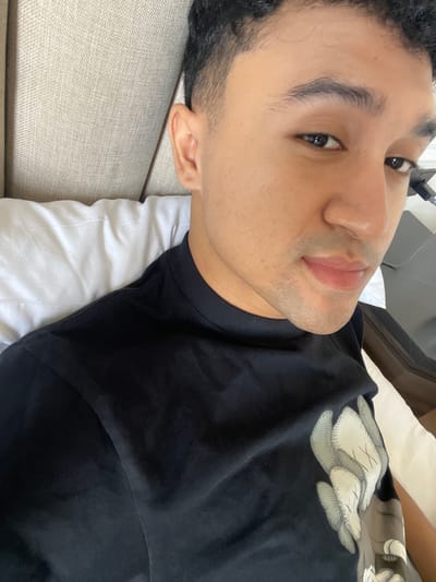 A young man with short, dark, curly hair and medium skin tone is depicted in Kayuambon, Indonesia, reclining against a beige, textured headboard. He is wearing a black crew-neck t-shirt adorned with a graphic featuring stylized light-colored shapes with 'X' marks on the right side. A white pillow is visible beneath his neck. He looks directly at the camera with a slight smile, revealing stubble on his chin and upper lip. A dark object, possibly furniture or electronics, is partially visible in the background to his right.