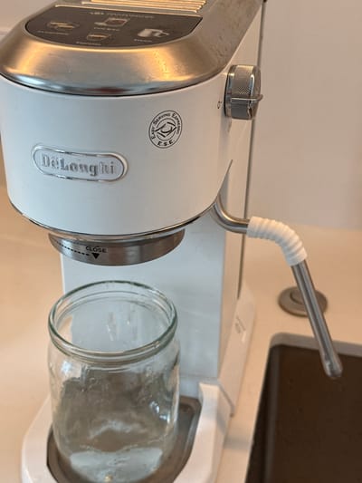 A close-up, eye-level, indoor shot shows a white De'Longhi espresso machine with a metal steam wand pointing downwards. A clear glass jar filled with a clear liquid sits beneath the steam wand. The machine has a silver control dial on its right side and a circular E.S.E. (Easy Serving Espresso) logo on its front. The machine is positioned on a white countertop, and a dark brown sink is partially visible to the right. The background is a plain, light-colored wall. There is no indication of the city of Гамово, Russia in the image.