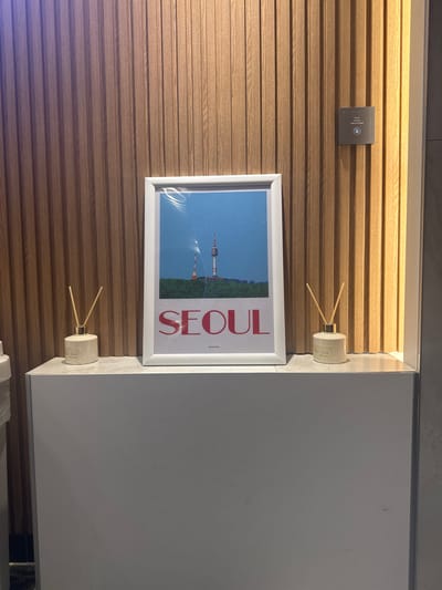 The scene depicts an indoor environment within a structure in 종로구, South Korea. A white-framed poster is centrally positioned on a light grey, counter-like surface. The poster features a depiction of the Namsan Tower and an adjacent smaller tower on a green, forested hill, set against a clear blue sky. Below the imagery, the word "SEOUL" is prominently displayed in large red capital letters, with a smaller, illegible word beneath it.
Flanking the poster, one on each side, are two cream-colored cylindrical reed diffusers with gold-colored necks, each containing multiple light brown wooden reeds and bearing small, illegible labels. The counter surface, which supports these items, appears to be the top of a larger, plain light grey structure.
The background consists of a vertically slatted wall constructed from light brown wood. Mounted on this wooden wall, to the right of the poster, is a dark grey rectangular control panel or sensor, featuring a circular blue light and minor, illegible text. No individuals are present in the image.