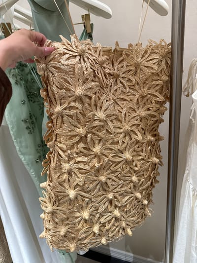 A hand is holding up a rectangular woven handbag with straw-like floral embellishments. The bag is hanging on a metal rod in what appears to be a closet or storage area with various garments. A green beaded garment is partially visible beneath the bag, along with other white or light-colored garments in the background. The location of this image is Sunrise, United States of America.