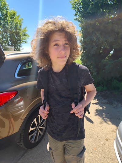 A fair-skinned adolescent with curly, light brown hair stands on a paved surface beside two cars, possibly in Chaponnay, France. The subject is wearing a patterned black t-shirt, olive green cargo shorts, and a black backpack. The first car is a light brown hatchback with its trunk open. The background shows trees and a bright blue sky with a sun glare. The subject is holding the straps of the backpack.
