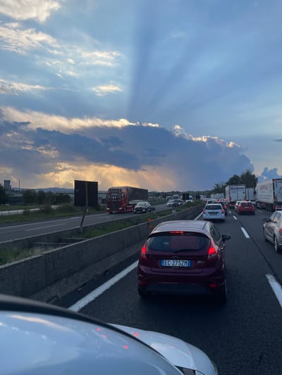 The image depicts a heavily congested multi-lane highway in the vicinity of Paliano, Italy. The sky above features prominent crepuscular rays with sunlight breaking through dark and light clouds, indicating either sunrise or sunset. In the immediate foreground, the white hood of the capturing vehicle is visible, showing reflections of red lights. Directly in front, a dark red Ford Fiesta hatchback is stationary in traffic, displaying Italian license plate `EC 275ZM` with its brake lights illuminated.

A concrete median barrier separates the vehicle's lanes from the opposing direction of travel. On the far left, a red semi-trailer truck is visible on the opposing highway lanes, followed by other vehicles. Further left, industrial-type structures and vegetation are present on the distant horizon, beneath a dark, rectangular signpost. The highway ahead is densely packed with stationary or slow-moving passenger cars and several white semi-trailer trucks, extending into the distant horizon. No individuals are distinctly visible.