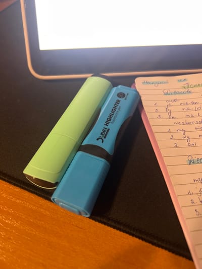 This image depicts a close-up, overhead view of a desk surface, likely in Skawina, Poland. The scene features two highlighters, one light green and one blue, lying next to a notepad. The notepad has pink edges and is filled with handwritten notes in blue ink, some of which are highlighted in yellow. A portion of a tablet or laptop screen, displaying a bright white, is visible in the upper left quadrant. The desk itself appears to be a black mousepad, with an orange surface partially visible in the bottom right. The lighting suggests an indoor environment, possibly during the day.