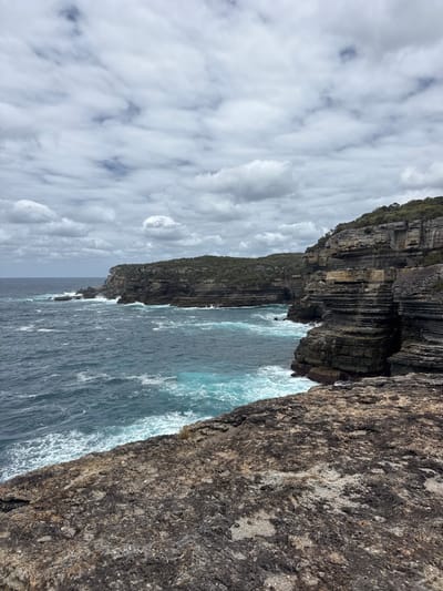 Jagged cliffs meet the ocean under a cloudy sky. The cliffside displays layered rock formations and sporadic vegetation. Turquoise water crashes against the base of the cliffs, creating white foamy waves. The foreground features a textured, dark brown rock face, contrasting with the distant horizon. The perspective is slightly angled, emphasizing the height and ruggedness of the coastal landscape.