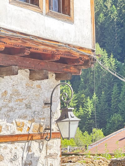 The image shows an exterior wall of a building in Shiroka Laka, Bulgaria. The wall is constructed of rough stone with sections of plaster. A wooden beam runs horizontally across the wall. A black metal lamp hangs from an ornate bracket attached to the wall. The lamp has a glass enclosure. Above the stone wall, there is a wooden eave supported by decorative wooden brackets. Overhead, wires are strung across the scene. In the background, a dense forest of green pine trees is visible. A portion of a red-tiled roof is also present in the lower right corner of the image. The lighting suggests it is daytime.
