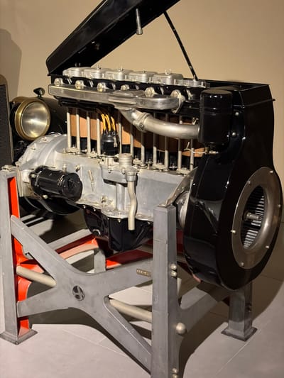 This media file captures a large, vintage internal combustion engine displayed on a stand. The engine is predominantly silver-grey metal, contrasted with black components and accents.
The main subject is a multi-cylinder engine, likely an inline configuration, visible with numerous exhaust pipes curving upwards and away from the block. On the side, an ignition system is visible with yellow/orange spark plug wires connected to the engine cylinders. A black, cylindrical starter motor is mounted near the bottom of the engine block.
A prominent feature is a large, glossy black housing on top of the engine, which appears to cover an overhead mechanism. This housing has a circular, metal-rimmed opening with internal vanes, possibly indicating a supercharger or a cooling fan assembly. Below this, chrome-like exhaust manifolds are visible, leading into larger pipes. Part of a black vehicle fender and a brass-rimmed headlight are visible in the lower left, indicating the engine is from an automobile.
The engine is mounted on a sturdy display stand made of grey metal, featuring bold, angular supports that form an X-shape at its base. The base of the stand also incorporates sections painted in a vibrant orange. The setting appears to be indoors, likely a museum or exhibition space, with a plain, light-colored wall serving as the background. The overall impression is one of showcasing engineering and automotive history.
There is no clearly legible text on the engine or stand, though some faint, unreadable markings appear on the grey engine block and a stylized symbol is present on the central hub of the X-shaped stand support. The lighting is artificial, suggesting an indoor environment.