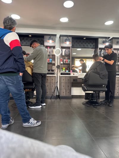 The scene depicts the interior of a barber shop in Krems an der Donau, Austria. The floor is covered with large, dark grey tiles. Four circular recessed lights are visible on the white ceiling, along with two dark-colored pendant lights positioned above barber stations.
In the immediate foreground on the left, a male subject with grey hair, wearing a navy blue jacket with red and white accents, blue jeans, and grey New Balance sneakers, stands with his back partially towards the camera.
Mid-ground left, a male barber with dark hair and glasses, dressed in a light olive-green collared shirt and black trousers, black sneakers, is actively cutting the hair of a seated client. The client, facing away from the camera, is draped in a yellow and brown striped barber cape. This station features a large mirror framed by dark wood-look cabinetry, with shelves displaying hair products.
Mid-ground right, a second male barber with dark hair, wearing a black polo shirt and black trousers, black sneakers, is also cutting the hair of a seated client. This client, facing away, is draped in a black barber cape. This station mirrors the one on the left, with a large mirror and dark wood-look cabinetry displaying products. A white ring light on a black tripod stands between the two main barber stations.
In the reflection of the mirror behind the right barber, a third male barber, wearing a dark shirt, is visible cutting the hair of a younger client (child) whose face is reflected. The overall environment suggests a modern, functional barbershop setup.