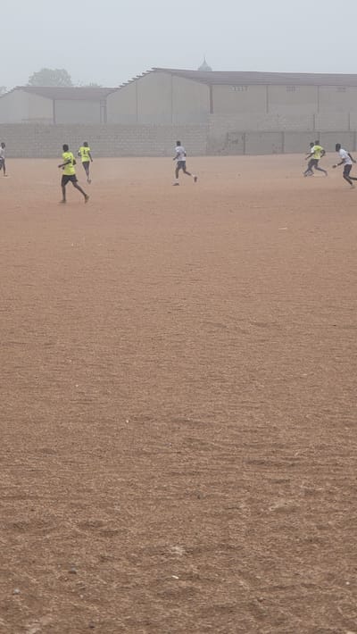 The image depicts a sports field in Jogana, Nigeria, with a building along one side. Several people are on the field, some running and some standing. The ground is dirt or sand. The weather is overcast or hazy.