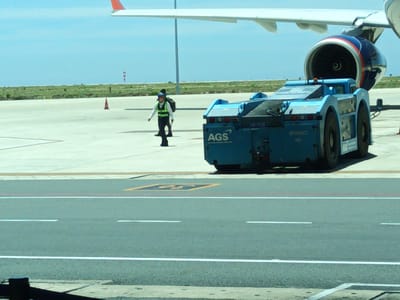 The image captures a scene at an airport in Bắc Cam Ranh, Vietnam, likely on the tarmac near a passenger airplane. In the foreground, a blue airport tug vehicle is positioned to the right of the frame, with the letters "AGS" visible on its side. The vehicle appears to be connected to a larger object, possibly an airplane's landing gear, although this is not clearly depicted. Behind the tug, a portion of an airplane's wing and engine are visible, suggesting the aircraft is either parked or being serviced. A figure in a high-visibility vest and dark pants is walking away from the viewer towards the airplane, carrying a bag over their shoulder. Another person, also in similar attire, is partially visible behind them. The environment is an open, paved area under a clear blue sky, with distant green fields and a few windsock-like structures in the background. The overall impression is of airport operations in progress on a sunny day.