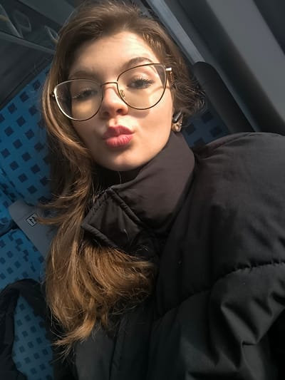 The image displays a close-up portrait of a female individual captured within a passenger vehicle, situated in the city of Kopanitsa, Bulgaria. The subject has light skin, long light-brown hair, and wears gold-framed cat-eye glasses, which exhibit reflections. Her lips are pursed, and she wears a black puffer jacket with a high collar over a dark garment. A black earbud is visible in her right ear. The background consists of blue fabric seats with a subtle lighter blue checkered pattern, indicative of public transport. A dark, rectangular window or panel is visible behind the subject's right side, and a light-colored object with a dark marking is partially visible on the seat to her lower left. Strong natural light illuminates the subject from the left.