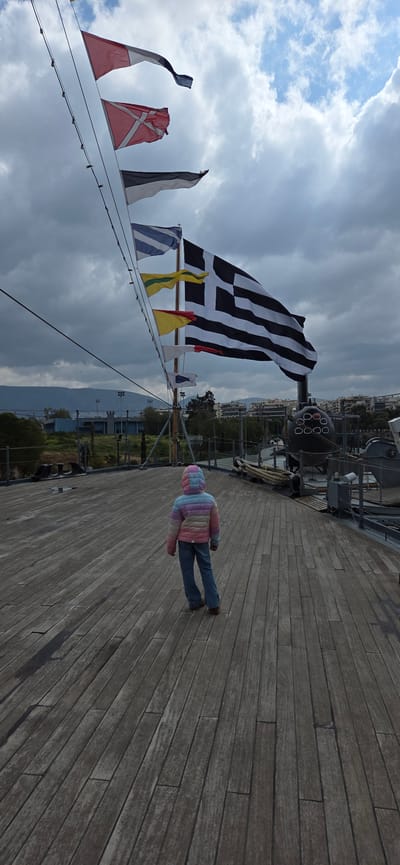 The scene depicts a single child standing on the weathered wooden deck of a ship, viewed from behind, at the Park of Maritime Tradition in Palaio Faliro, Greece. The child wears a multi-colored striped hooded puffer jacket (displaying pink, blue, yellow, and white segments) and blue jeans, facing towards the bow of the vessel.

A series of flags are displayed overhead, extending upwards and diagonally from a mast and rigging on the left against a cloudy sky with visible patches of blue. From top to bottom, these include a white over red flag (Hotel), a red flag with a white Scandinavian cross (Flag of Denmark), a black over white flag (November), a blue and white striped flag with a white cross in the canton (Greek Naval Jack), a yellow flag with a horizontal green stripe (Lima), a yellow flag with a red square on the hoist (Juliet), and a white flag with a red square on the hoist (Sierra). A large flag, rendered in black and white tones, depicting nine horizontal stripes and a white cross on a black square canton (Greek National Flag), is prominently displayed to the right of the signal flags.

On the deck to the right, ship components such as metal railings, coiled ropes, and a dark cylindrical object with multiple circular openings, possibly a torpedo tube array, are visible. The background shows an urban landscape featuring buildings, trees, and distant hills or mountains. The wooden deck planks are aged, displaying wear and varying tones of grey and brown.