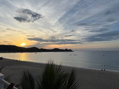 The image depicts a sunset scene at a beach in Song Cau, Vietnam. The sky is filled with a mix of clouds, with streaks of orange and yellow near the horizon where the sun is setting. The sea is calm, reflecting the colors of the sky. The beach is sandy, with a few people walking along the shoreline. Palm fronds are visible in the foreground, adding a tropical element to the scene. Several boats are dotted in the water and along the coast. In the distance, hills form a silhouette against the sky. The overall atmosphere is serene and peaceful.